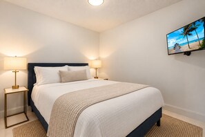 A neatly made bed with a dark headboard, two bedside lamps, and a wall-mounted TV displaying a beach scene. - Restoration Hardware PGA National Condo w/ 180° Golf Course Views (Palm Beach Gardens)