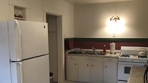 Keittiössä on valkoiset kaapit, jääkaappi, liesi ja pesuallas. Lattialla on matto ja etualalla tuoli.