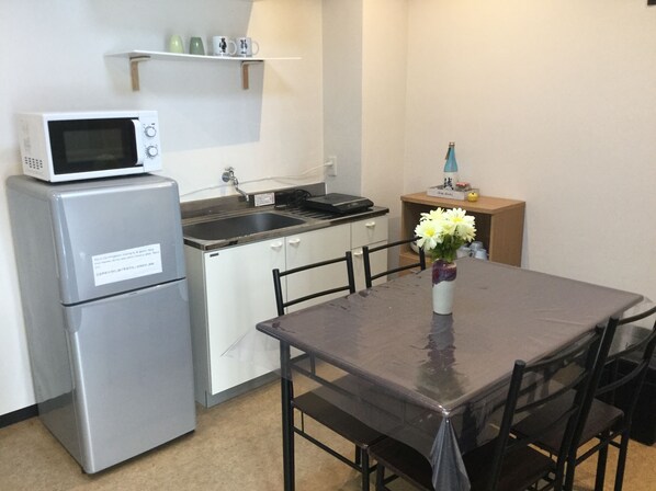 A compact kitchenette with a microwave, refrigerator, sink, and a small shelf with cups. There's a dining table with chairs and a vase with flowers.