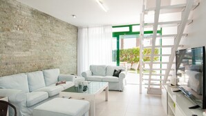 Ein modernes Wohnzimmer mit Backsteinwand, zwei helle Sofas, einem Glastisch, einer weißen TV-Konsole und einer Treppe, die zu einem Außenbereich führt.
