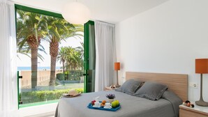 Ein modernes Schlafzimmer mit einem großen Bett, einem Nachttisch, einer Lampe und Blick auf Palmen und einen Strand durch eine Glastür.