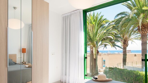 Ein modernes Hotelzimmer mit großem Fenster und Blick auf Palmen und den Strand.