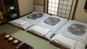 A traditional Japanese room with tatami mats, two tatami beds with floral bedding, a wooden table with a remote control and a book, and a TV on a shelf. - Nihonkan (Kyoto)