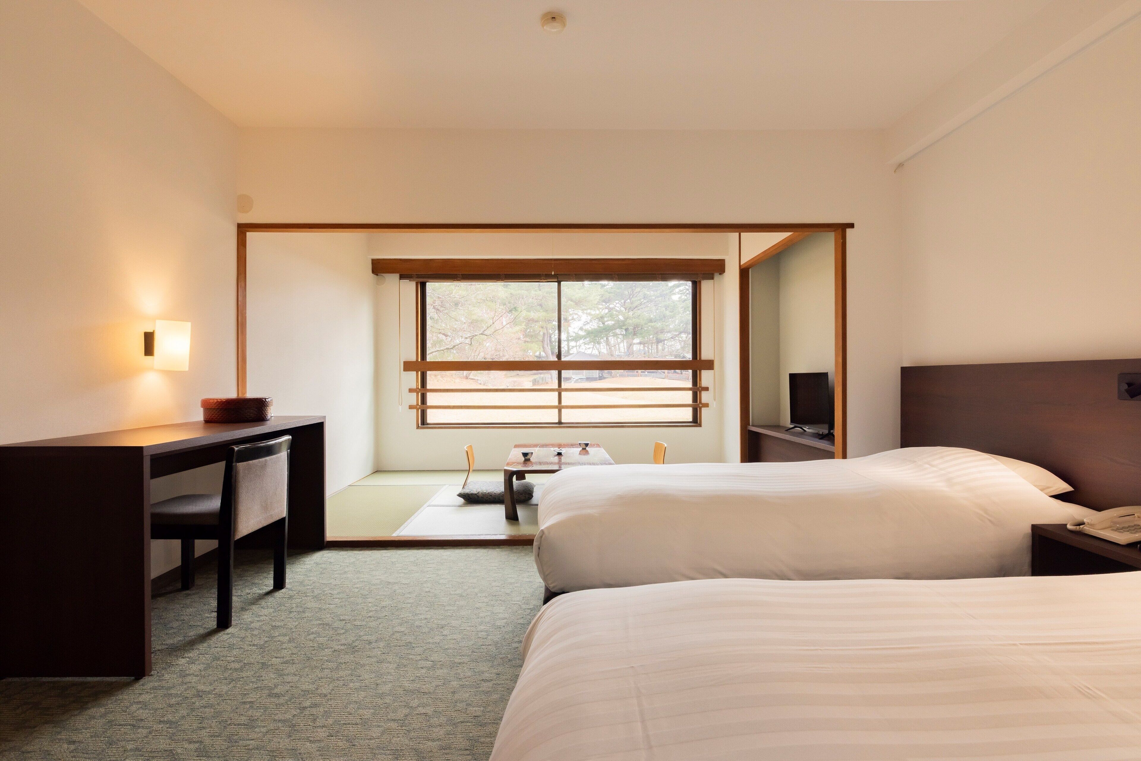 A hotel room with two beds, a desk, a chair, a small table, a mirror, and a window with blinds.