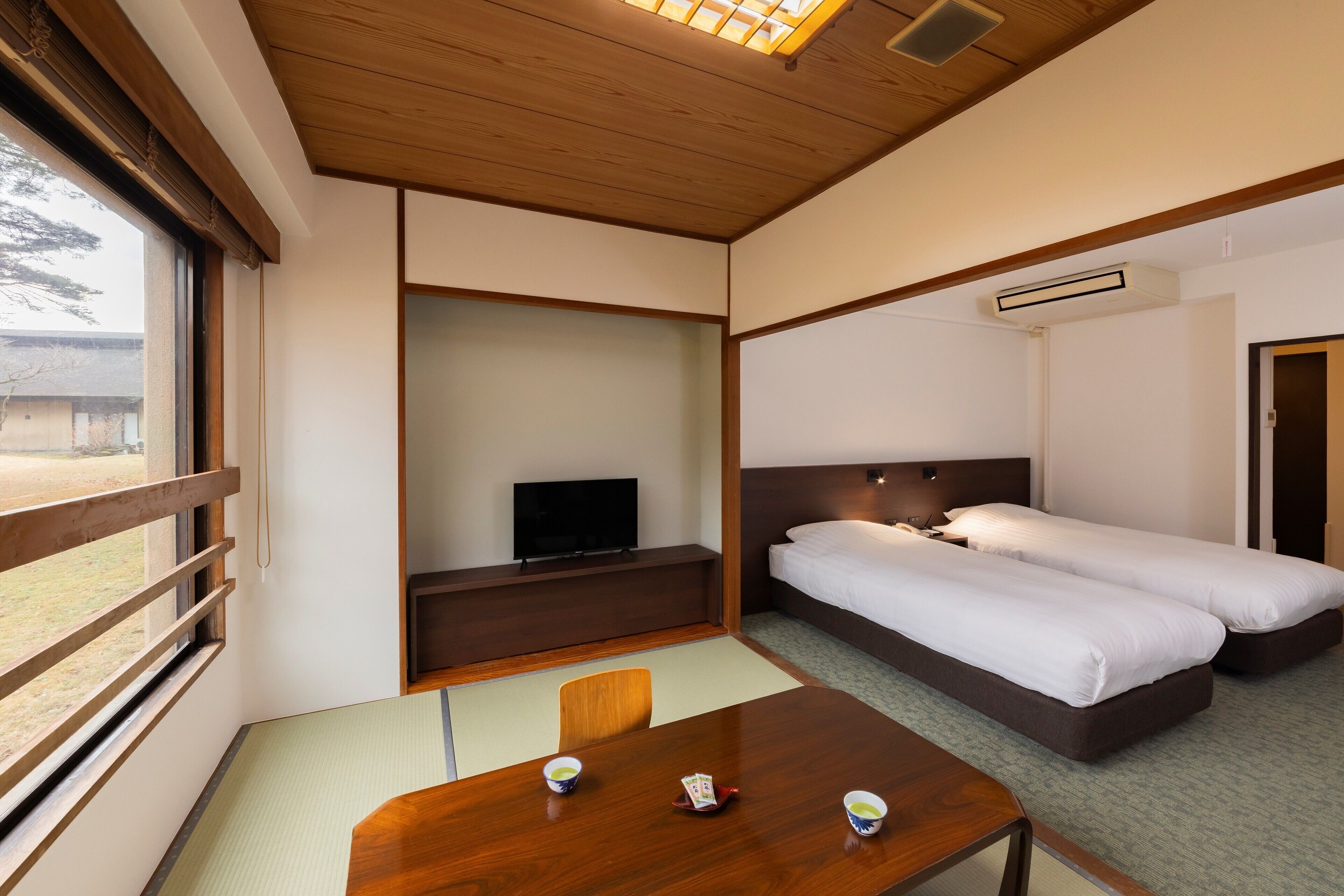 Habitación de hotel con techo de madera, un televisor en un estante, dos camas, una mesa con tazas y vistas al exterior.