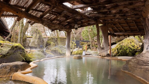 Un onsen japonés tradicional con una estructura de madera sobre él, rodeado de rocas y musgo.