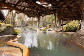 A traditional Japanese onsen with a wooden structure overhead, surrounded by rocks and moss. - Aso Baien Spa Resort (Minamiaso)