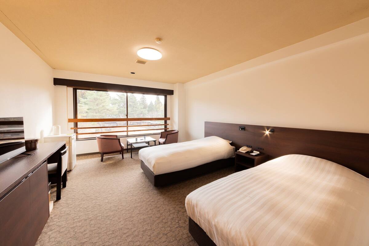 Une chambre d’hôtel avec deux lits, un bureau, une chaise, une fenêtre donnant sur des arbres et une table de chevet avec une lampe.
