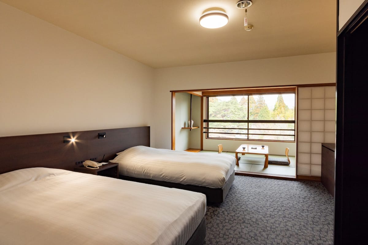 Une chambre d’hôtel avec deux lits, une petite table, une chaise, une table de chevet avec un téléphone et une grande fenêtre donnant sur des arbres.