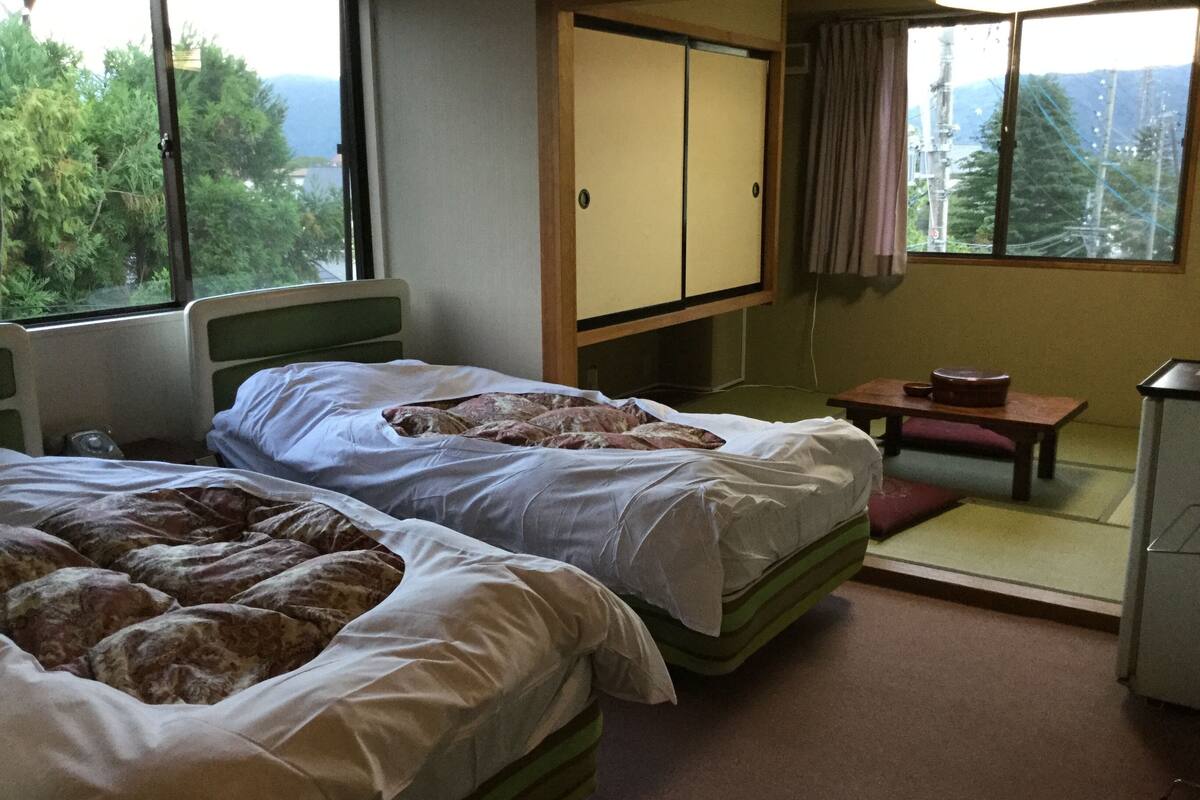 A hotel room with two beds, a small table, a cabinet, and a view of the outdoors.