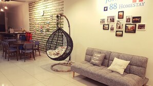 Una sala moderna con una silla colgante, un sofá gris con cojines decorativos, un comedor con sillas, y una pared decorada con cuadros y un letrero que dice "JONKER 88 Homestay".