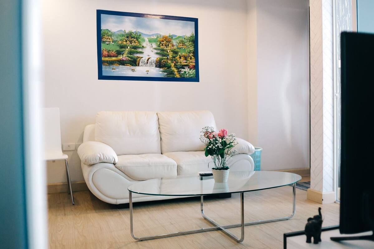 A modern living room with a white sofa, a glass coffee table, and a painting of a landscape on the wall.