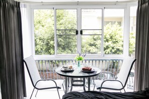 Ein Zimmer mit einem runden Tisch für zwei Personen, einer Blumenvase und Blick auf Bäume durch große Fenster.
