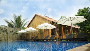 A swimming pool with lounge chairs and umbrellas, wooden buildings, and palm trees in the background.