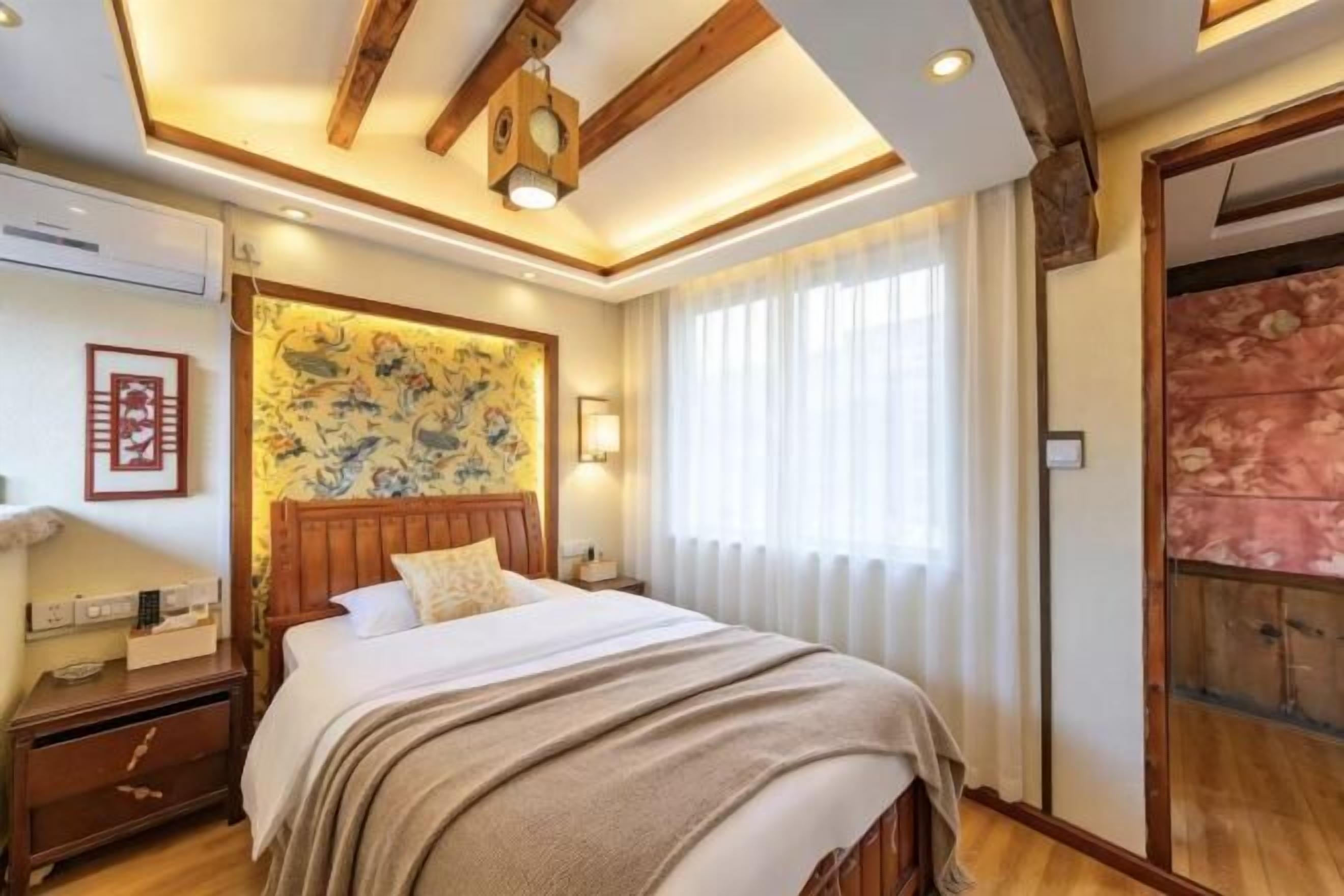 A bedroom with a wooden headboard, a bed with a white comforter, a nightstand, a wall-mounted lamp, and a large window with sheer curtains.