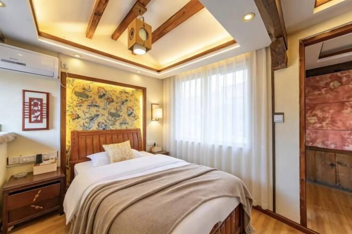 Une chambre à coucher avec une tête de lit en bois, un lit recouvert d’une couette blanche, une table de chevet, une lampe fixée au mur et une grande fenêtre ornée de rideaux légers.