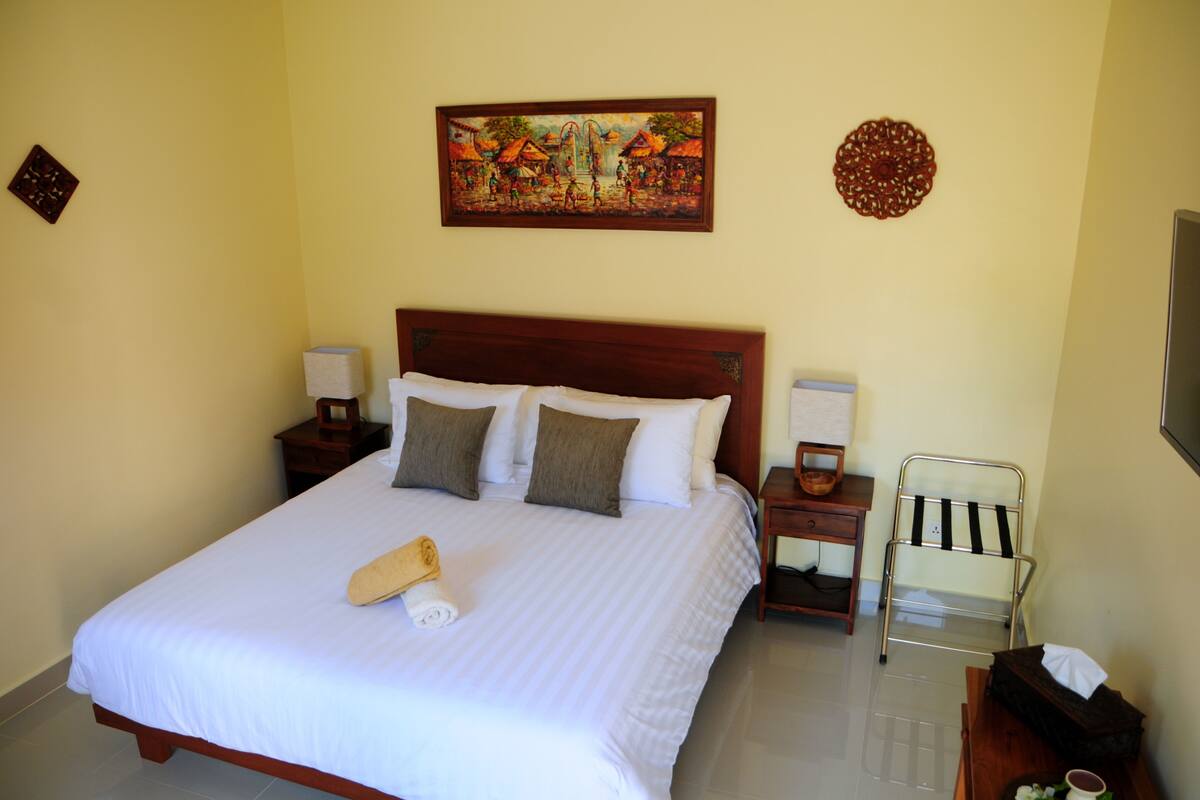 A neatly made bed with pillows, a bedside table with a lamp, a chair, and a wall painting.