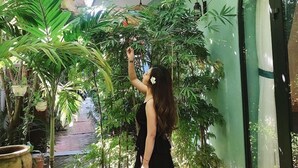 Una donna con un abito nero che allunga la mano verso una pianta in un giardino interno con pareti verdi, varie altre piante e un lampadario.