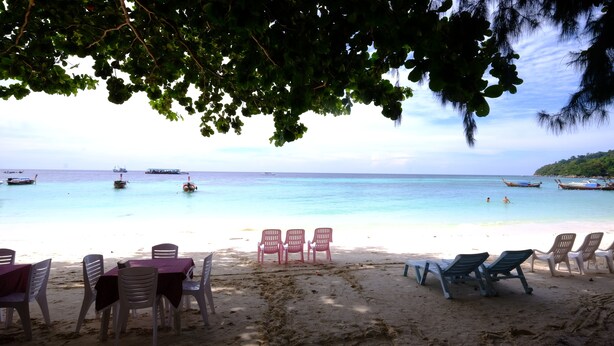 A beach with clear turquoise water, several boats, and a row of plastic chairs and tables under a tree. - Seaside Resort Koh Lipe (Koh Lipe)
