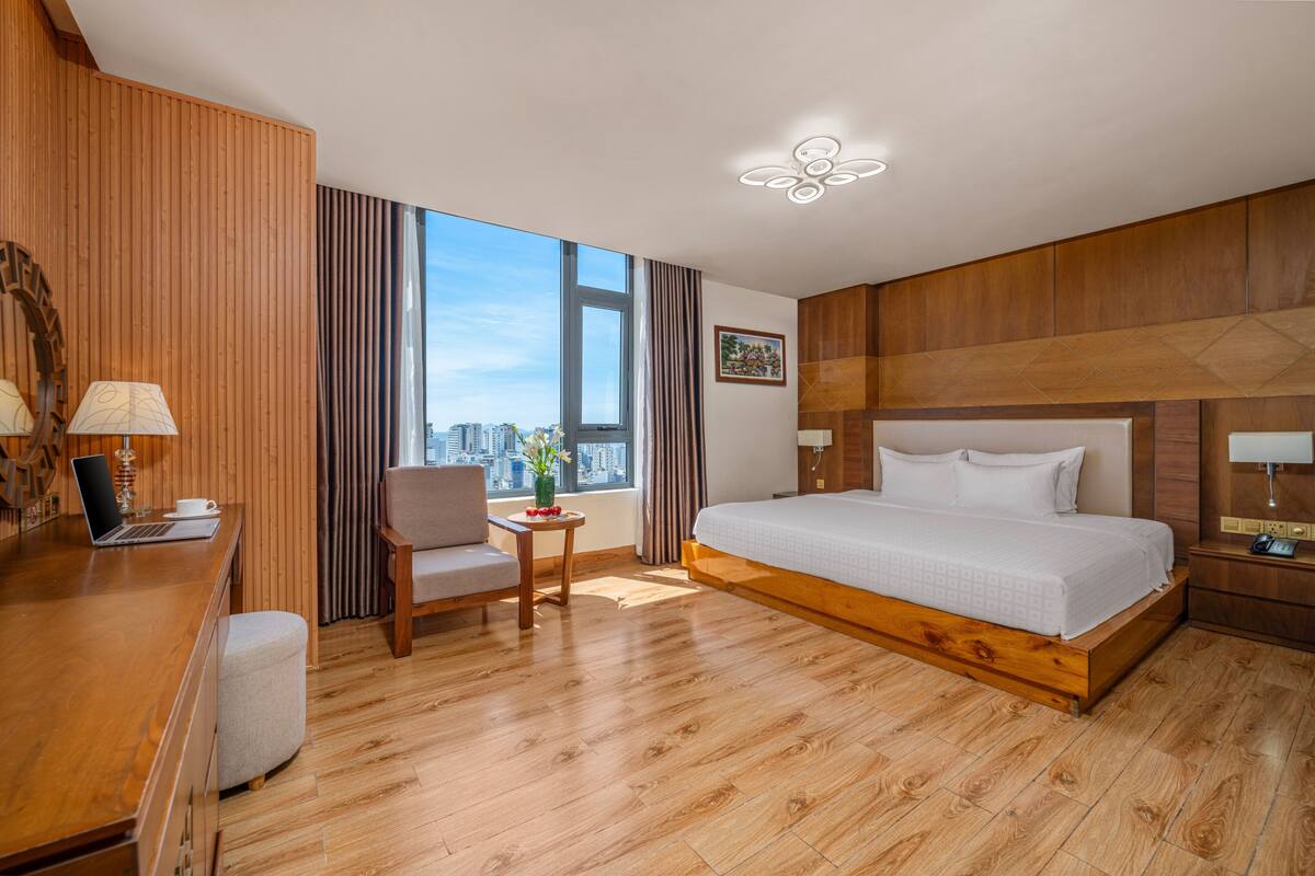 A hotel room with a wooden floor, a bed, a desk with a laptop, a chair, a small table with a vase, and a large window with a city view.