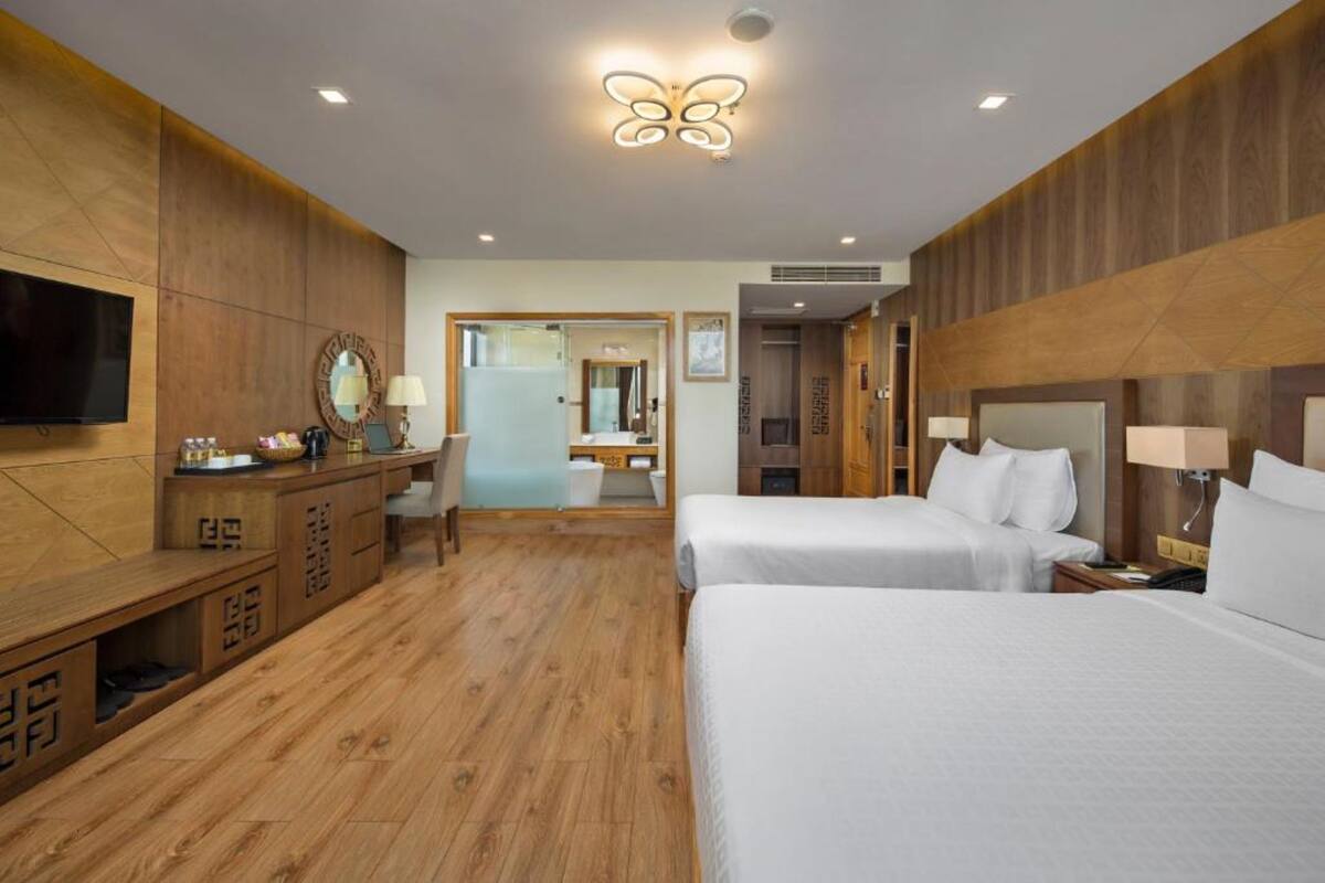 A hotel room with wooden paneling, a TV, a desk with a mirror, two beds, and a bathroom visible through an open door.