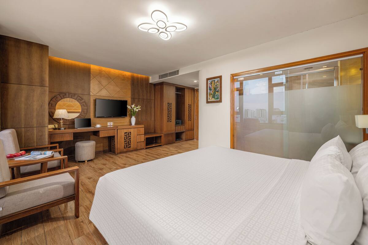 A hotel room with a large bed, a desk with a computer, a TV, and a city view through the glass door.