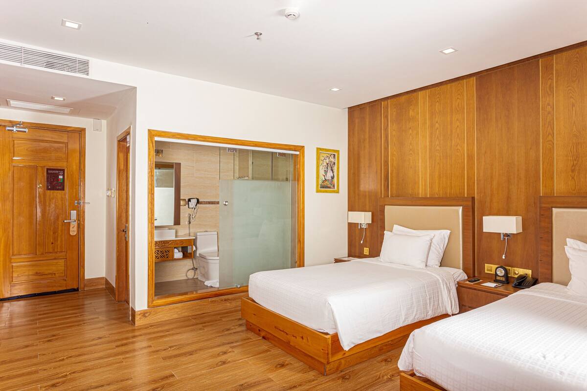 A hotel room with two beds, wooden flooring, a wooden wardrobe, a bathroom with a glass shower enclosure, and a wooden panel wall.