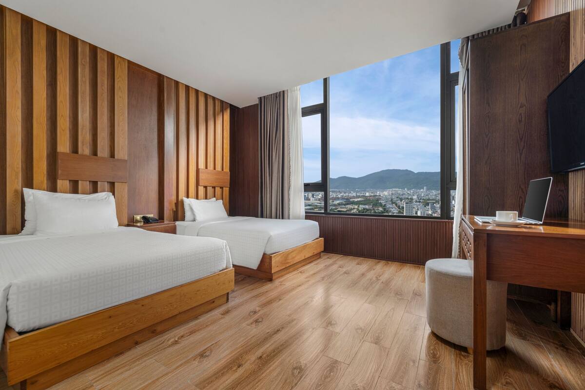 A hotel room with two beds, a wooden floor, a desk with a laptop, and a large window offering a city view.