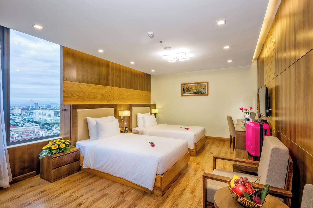 A hotel room with two beds, a wooden headboard, a desk with a chair, a TV, and a window with a city view.