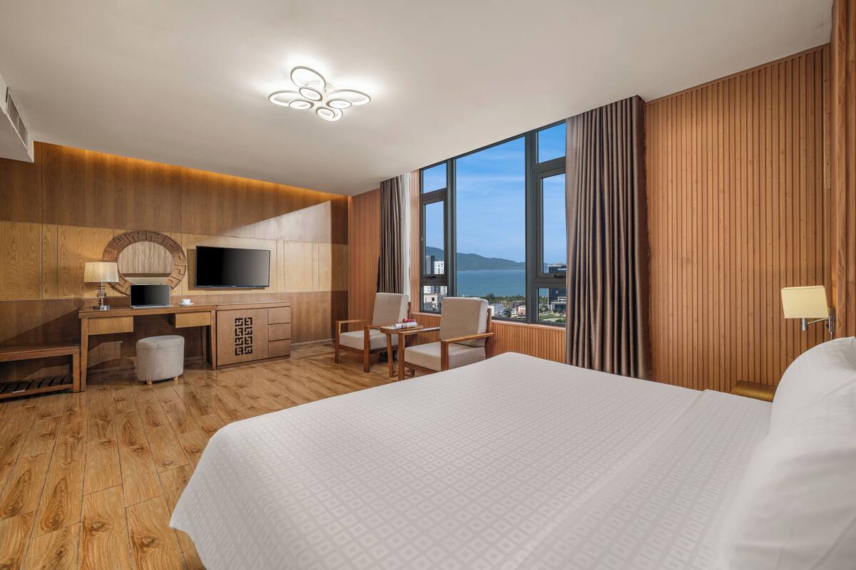 A hotel room with a large bed, a desk with a computer, a chair, and a TV. The room has wooden paneling and large windows with a view of the city.
