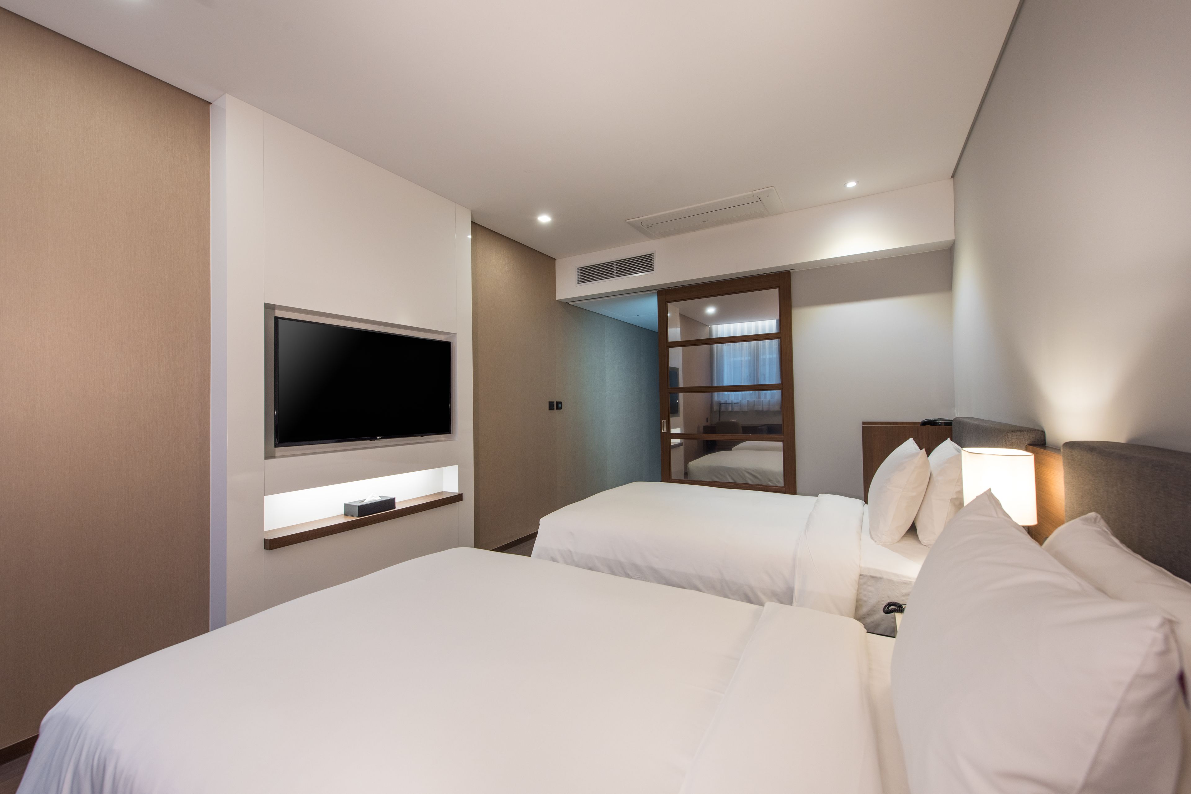 A hotel room with two beds, a flat-screen TV mounted on the wall, and a bedside table with a lamp.