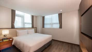 A hotel room with a large bed, a nightstand with a lamp, a flat-screen TV mounted on the wall, and two windows with curtains.