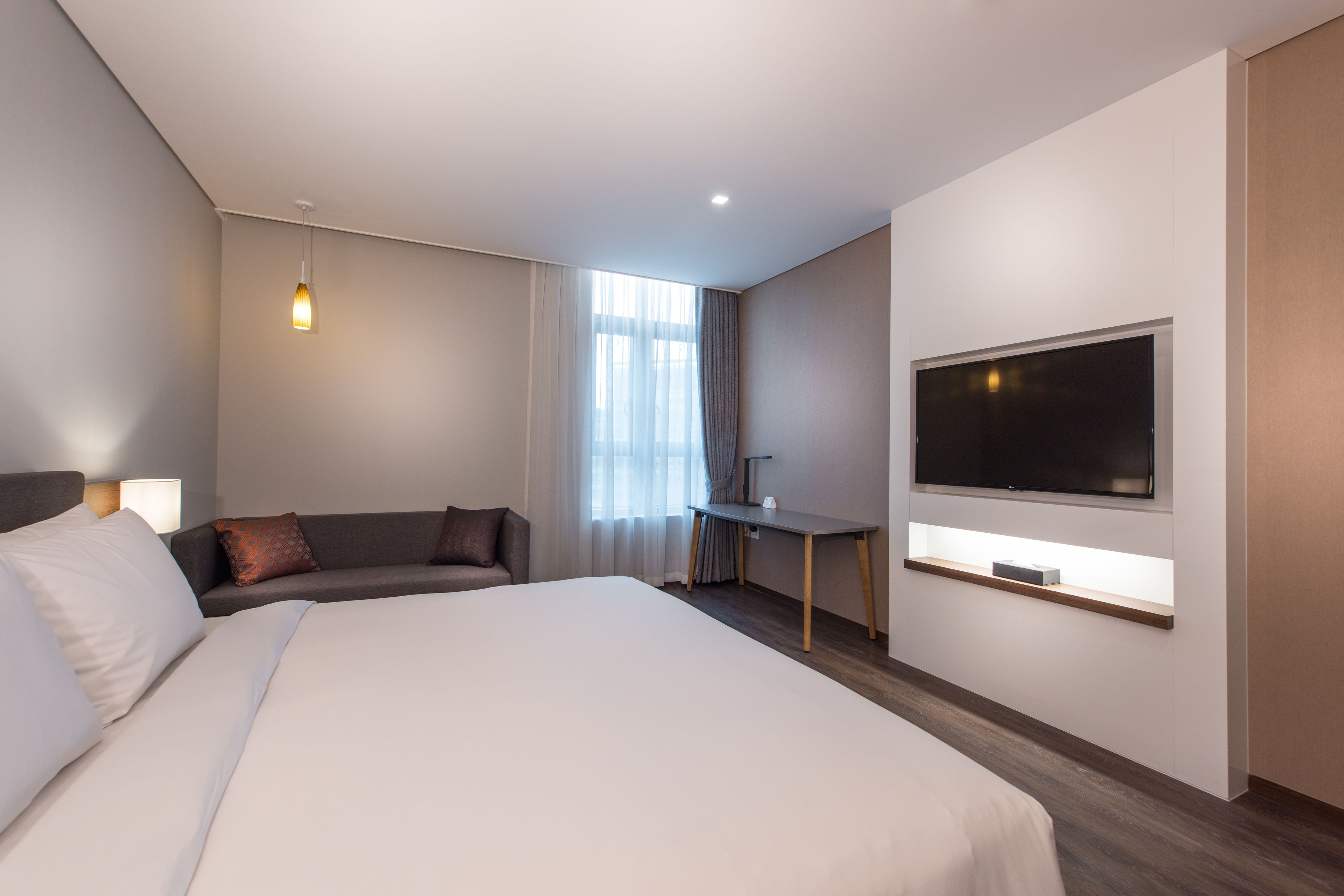 Ein modernes Hotelzimmer mit einem großen Bett, einer Sitzecke mit einer Couch und Sesseln, einem Schreibtisch mit einem Flachbildfernseher und einem großen Fenster mit Vorhängen.