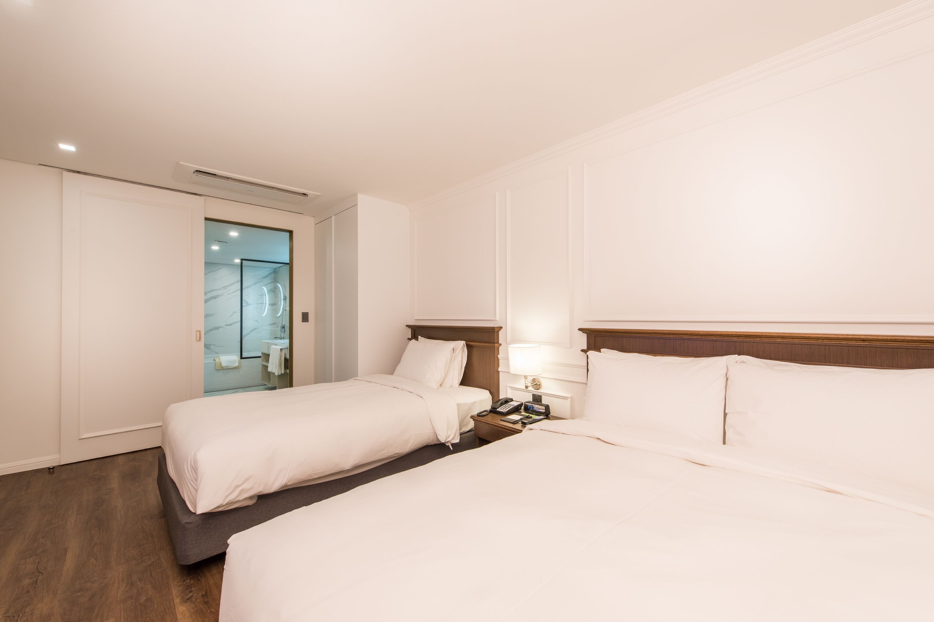 A hotel room with two beds, a wooden headboard, a bedside table with a lamp, and a glass shower enclosure.