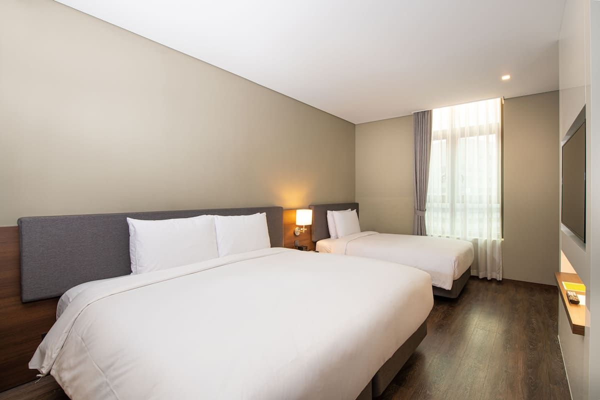 A hotel room with two beds, a wooden headboard, a nightstand with a lamp, a window with curtains, and a flat-screen TV mounted on the wall.