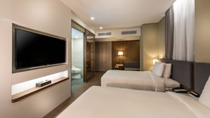 Une chambre d’hôtel moderne dotée d’un grand téléviseur à écran plat fixé au mur, de deux lits avec des têtes de lit, d’une table de chevet, d’une lampe et d’un dressing avec une porte coulissante.