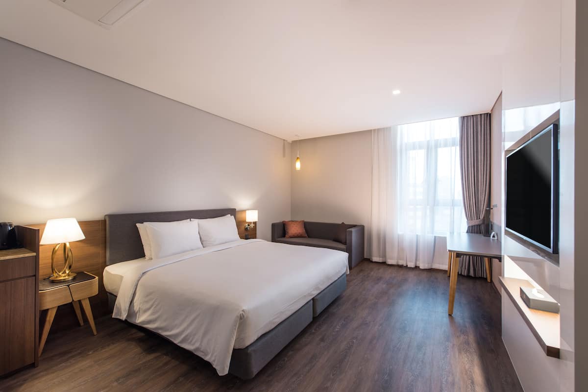 A modern hotel room with a large bed, bedside tables with lamps, a sofa, a TV, and a large window with curtains.