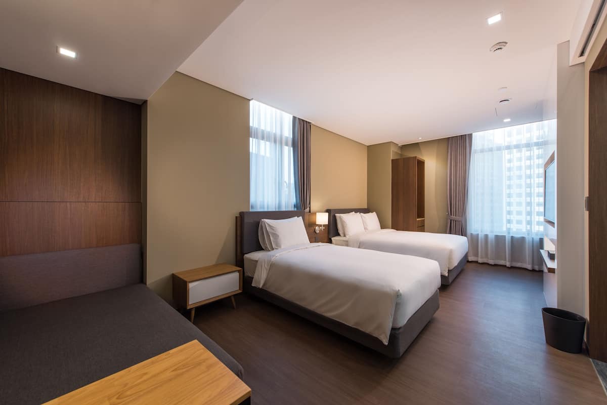 A modern hotel room with two beds, a sofa, a desk, and a large window with curtains.