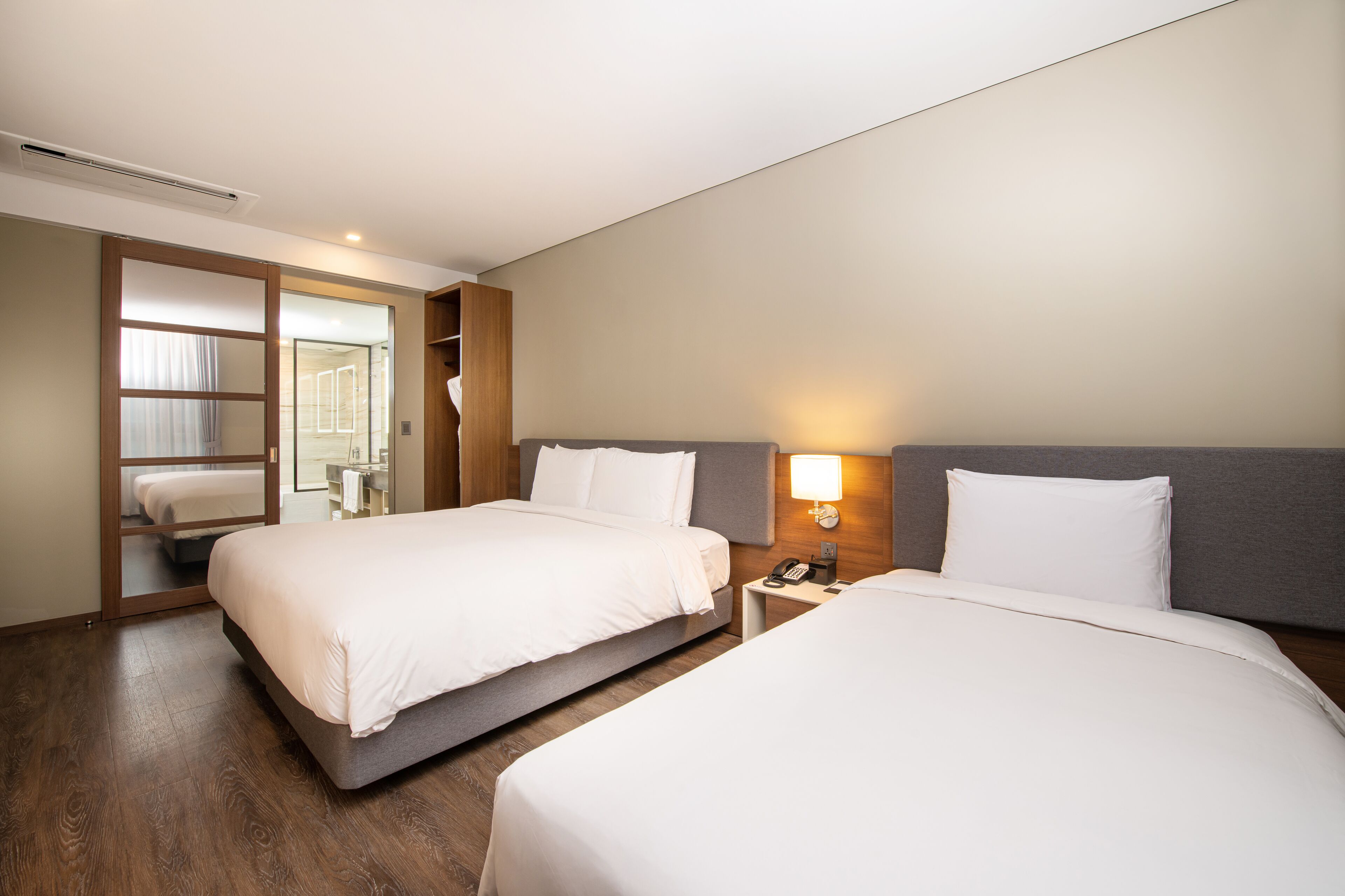 Camera d'albergo con due letti, un comodino con una lampada e una porta scorrevole che dà su un bagno.