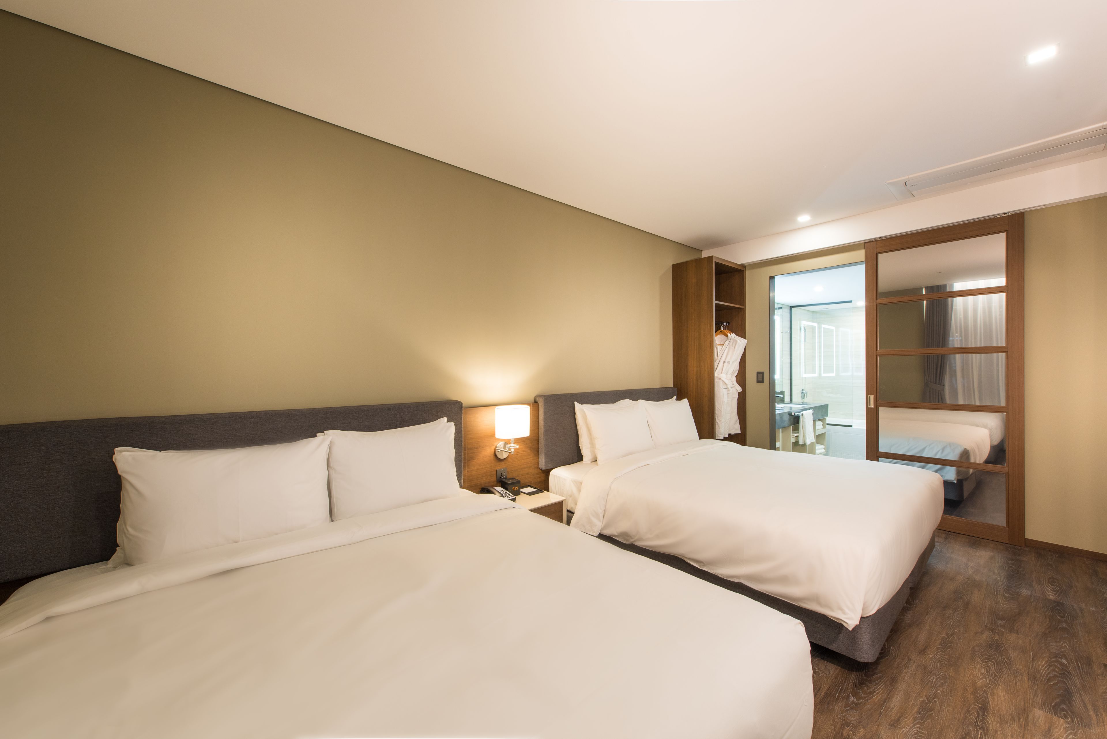 A hotel room with two beds, a wooden headboard, a nightstand with a lamp, and a sliding glass door leading to a bathroom.