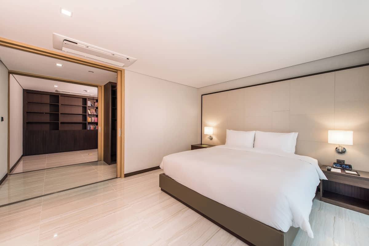 A modern hotel room with a large bed, a nightstand with a lamp, a built-in wardrobe, and a ceiling-mounted air conditioner.
