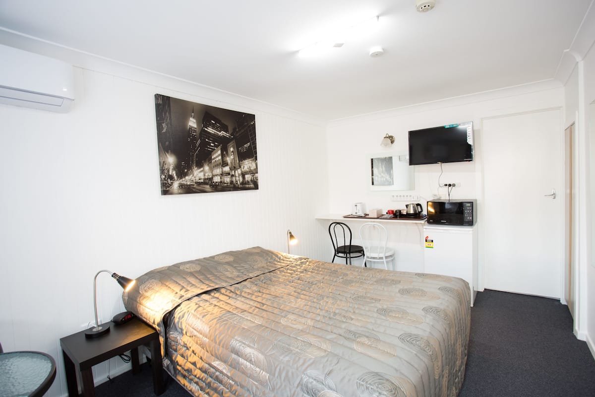 A hotel room with a bed, a desk with a computer, a chair, a flat-screen TV, and a wall-mounted air conditioner.