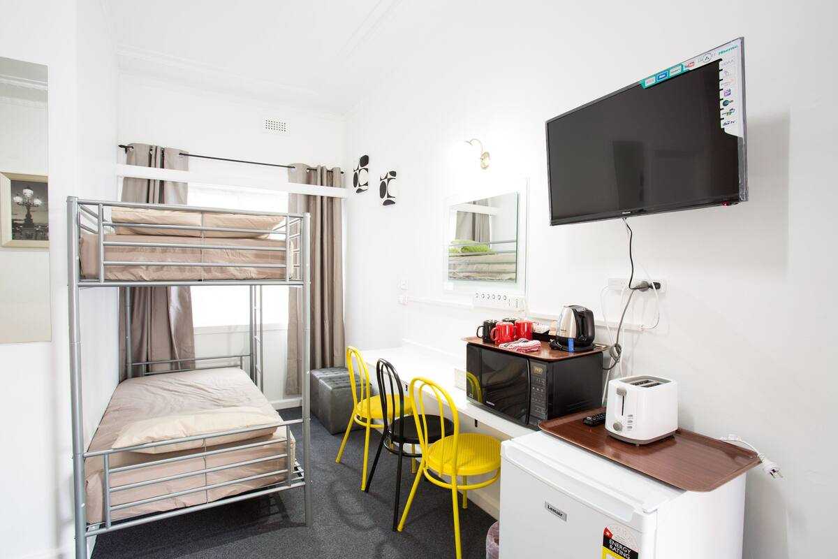 A compact hotel room with a bunk bed, a desk with a microwave, a toaster, and a television.