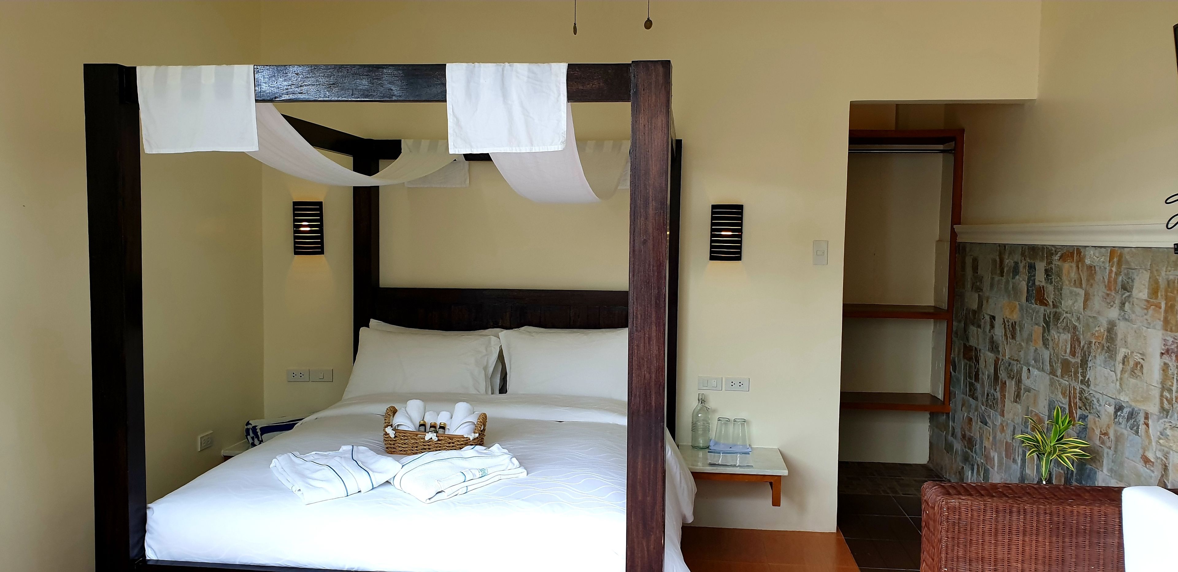 A four-poster bed with a basket of toiletries, a bedside table with glasses and a bottle, and a wicker chair with a plant.