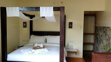 A four-poster bed with a basket of toiletries, a bedside table with glasses and a bottle, and a wicker chair with a plant.