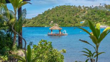 A tranquil beach scene with a thatched-roof hut on stilts in the water, surrounded by lush greenery and a clear blue sky.