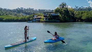 Two people paddleboarding on a calm lake with a house and lush greenery in the background.