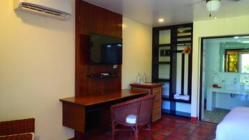 A hotel room with a wooden desk, a television mounted on the wall, a wicker chair, and a bathroom visible through an open door.