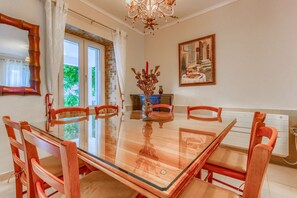 A dining room with a glass-top table, wooden chairs, a chandelier, a painting, and a window with curtains.