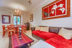 A living room with a red sofa, a dining table with chairs, a chandelier, and a large floral painting on the wall.
